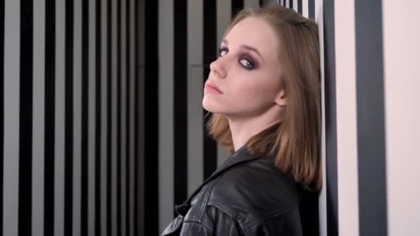 Junge hübsche Frauen mit schwerem Make-up in schwarzer Jacke an der Wand lehnend, sich umdrehend und in die Kamera blickend, gestreifter Wandhintergrund — Stockvideo