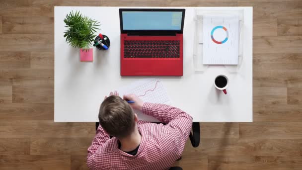 Jonge man in glazen werken, schrijven op papier, topshot, zittend achter bureau met koffie en laptop — Stockvideo