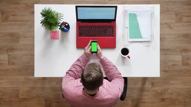 Jovem usando telefone com chave chroma, topshot, sentado atrás da mesa com laptop e café — Vídeo de Stock