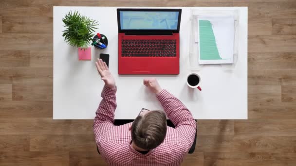 Joven con gafas hablando con alguien a través de laptop, topshot, sentado detrás del escritorio con café y documentos — Vídeos de Stock