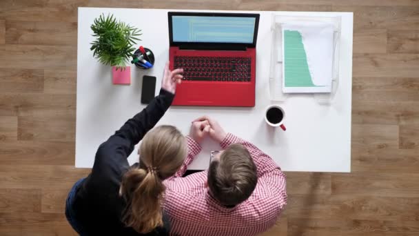 Ung man i glasögon pratar med kvinnor över laptop, topshot, sitter bakom skrivbord med kaffe och dokument — Stockvideo