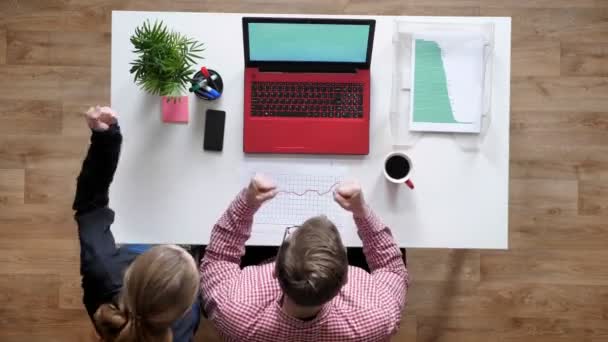 Ung man i glasögon och kvinna som tittar i laptop, jublande och ger hög fem, topshot, skrivbord med kaffe och dokument — Stockvideo