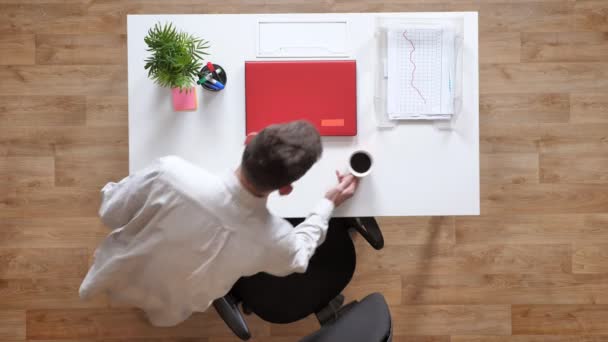 Junger Mann geht mit Kaffee rein, setzt sich und öffnet Laptop, topshot, sitzt hinter Schreibtisch — Stockvideo