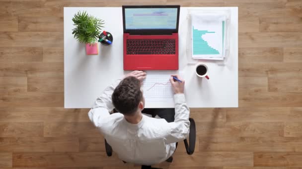 Jonge man in een wit overhemd schrijven op papier, topshot, zittend achter tafel met laptop en koffie — Stockvideo