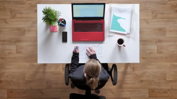 Молода жінка з хвіст записує на аркуші паперу і сидить за столом з ноутбуком і кавою, tophot — стокове відео