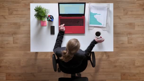 Jonge vrouw met een paardenstaart typen op laptop en koffie drinken, zitten achter de tabel met documenten, topshot — Stockvideo