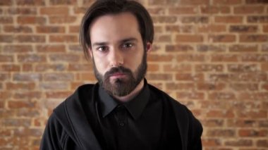 Genç siyah sakallı adam kameraya, tuğla arka plan izlerken sakin