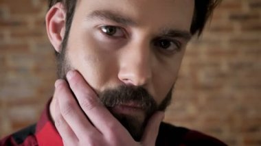 Genç çekici ciddi adam kameraya, tuğla arka plan izlerken onun sakalı dokunuyor