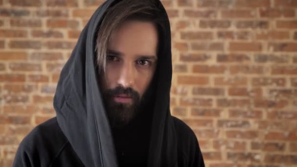 Young interesting man with beard in hood is watching at camera, goes away, brick background — Stock Video