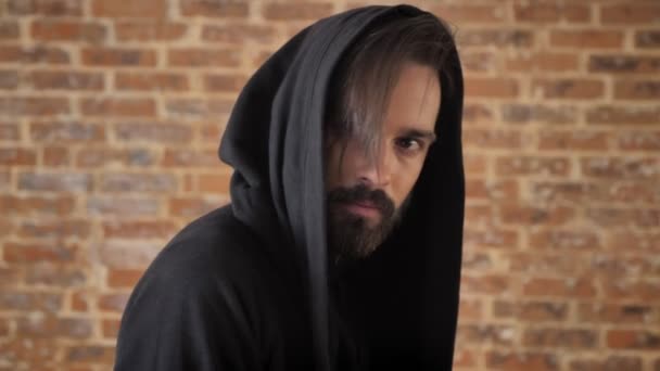 Young shocked man with beard in hood turns to camera, watching at camera, brick background — Stock Video