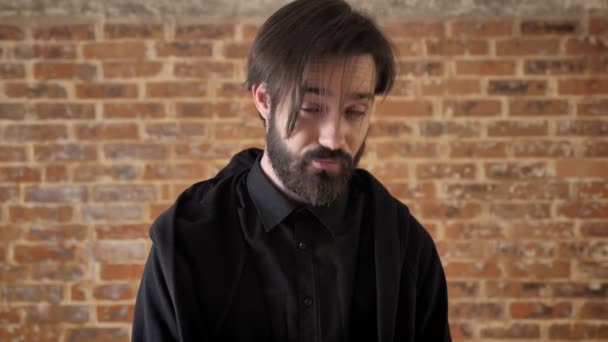 Young indifferent guy raises eyebrow, watching at camera, brick background — Stock Video