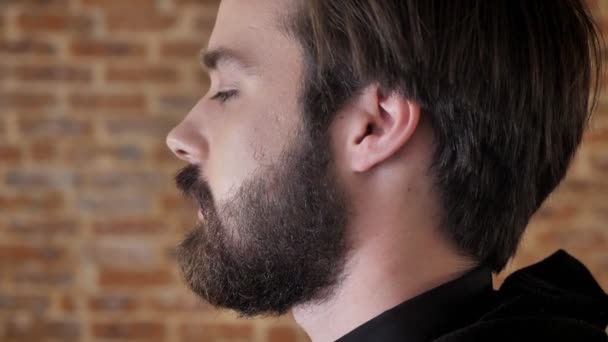 Молодой привлекательный мужчина с бородой поворачивает голову, наблюдает за камерой, улыбается, флиртует концепция, кирпичный фон, вид сбоку — стоковое видео
