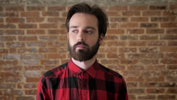 Jeune homme beau avec la barbe regarde à la caméra, fond de brique — Video