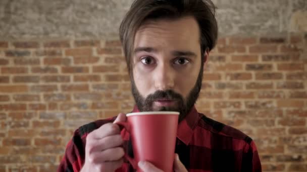 Молодой привлекательный мужчина с бородой пьет чай, смотрит на камеру, закусывает. — стоковое видео