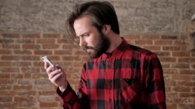 Sakallı genç yakışıklı adam onun smartphone, comunication kavramı, tuğla arka plan mesajları okur