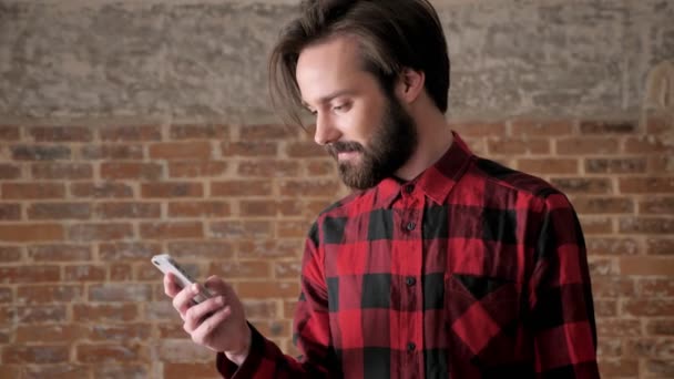Молодий красивий чоловік з бородою дивиться фотографії на свій смартфон, концепція спілкування, цегляний фон — стокове відео