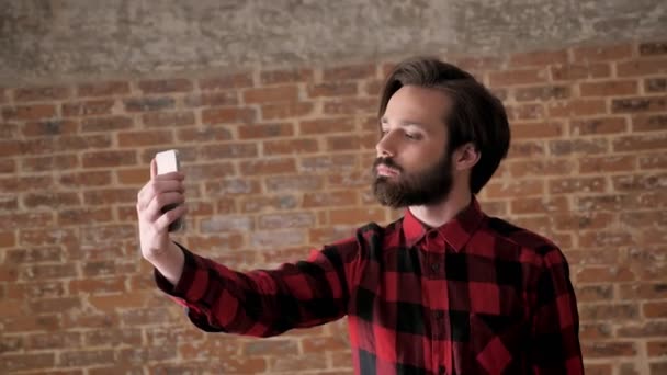 Joven hombre guapo con barba hacer selfie en su teléfono inteligente, concepto de comunicación, fondo de ladrillo — Vídeos de Stock