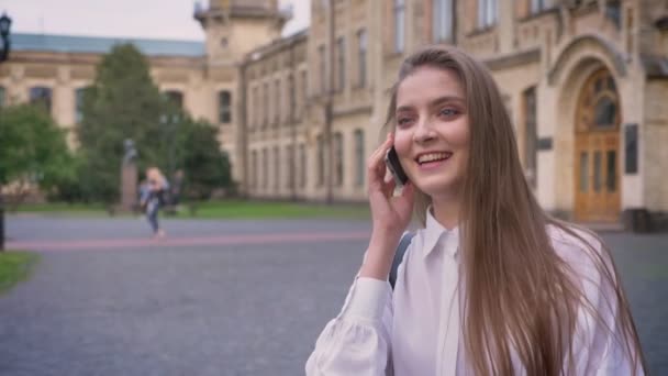 Menina feliz bonita nova está falando no telefone e indo da universidade durante o dia no verão, concepção de comunicação — Vídeo de Stock
