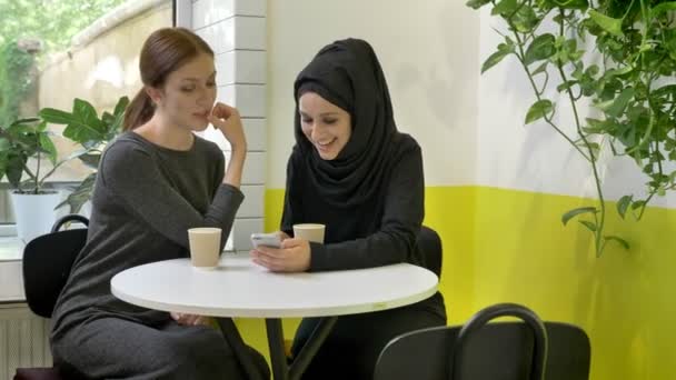 Bir tanesi telefon arayan ve konuşurken, hijab, kafede oturan iki genç güzel Müslüman kadının, gülüyor — Stok video