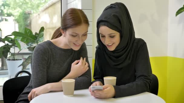 Two young womans sitting in cafe, one of them muslim woman in hijab, looking at phone and laughing, looking in camera — Stock Video