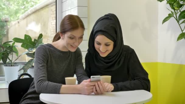 Dos mujeres jóvenes sentadas en la cafetería, una de ellas musulmana en hiyab, mirando el teléfono y mirando a cámara, sonriendo — Vídeos de Stock