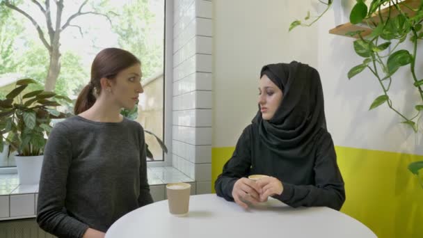 Zwei junge schöne Frauen sitzen im Café, eine davon Muslimin im Hidschab, die einer anderen Frau etwas erzählt — Stockvideo