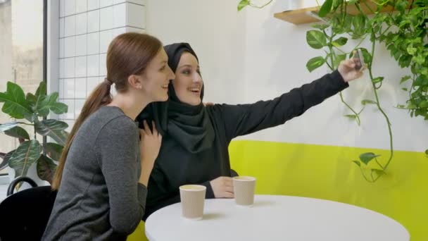 Дві молоді красиві жінки сидять в кафе, одна з них мусульманка в хіджабі, приймаючи селфі з телефоном і дивлячись по телефону — стокове відео