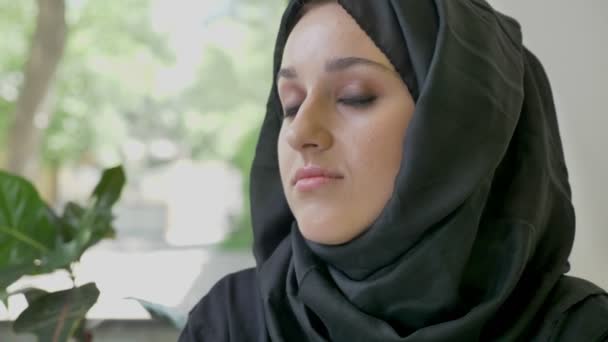 Retrato de una joven musulmana en hijab sentada en la cafetería, mirando directamente a la cámara y pensando, preocupada — Vídeos de Stock