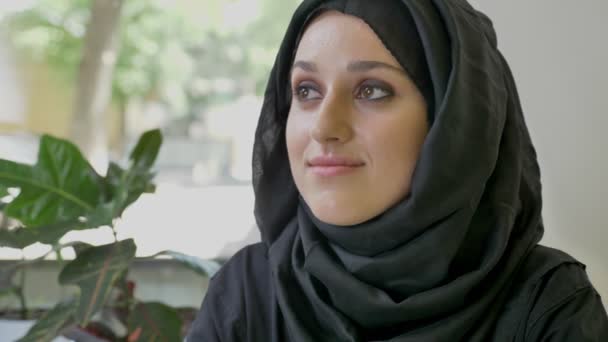 Портрет молодой красивой мусульманки в хиджабе, сидящей в кафе, смотрящей вперед, а затем в камеру, улыбающейся — стоковое видео