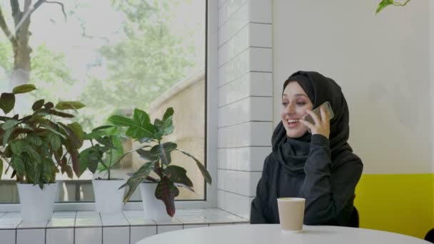 Joven mujer bastante musulmana en hijab hablando por teléfono y sonriendo, sentado en la cafetería, alegre — Vídeos de Stock