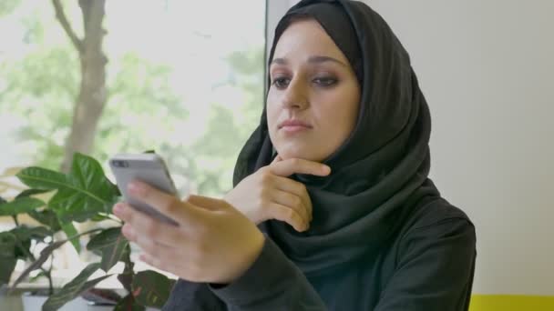 Porträtt av ung ganska muslimsk kvinna i hijab sitter i café, håller telefonen och ser fram emot, berörda och bestäms — Stockvideo