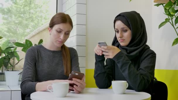 Dos mujeres hermosas jóvenes sentadas en la cafetería y escribiendo por teléfono, una de ellas musulmana en hiyab, decidida y concentrada — Vídeos de Stock