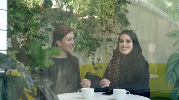 Две молодые женщины сидят в кафе, одна из них мусульманка в хиджабе, разговаривает и смеется, вид со стороны — стоковое видео