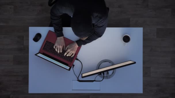 Ung man i svart hoodie att skriva på laptop och tittar på bildskärm under natten, sitter bakom vita skrivbord med kaffe, topp skott — Stockvideo