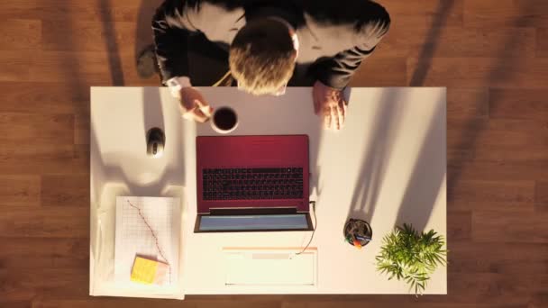 Jovem de óculos e fato, bebendo café e digitando no laptop, sentado atrás da mesa, tiro superior — Vídeo de Stock