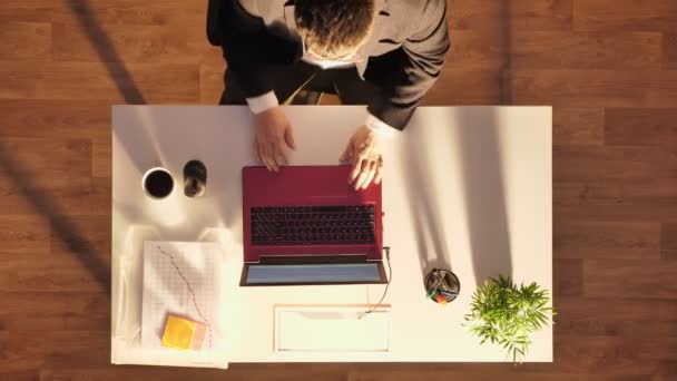 Молодой человек в очках и костюме сидит за столом с ноутбуком и кофе, уходя, верхний выстрел — стоковое видео