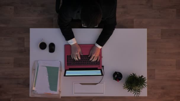 Jeune homme en lunettes et costume assis derrière le bureau, dactylographiant sur ordinateur portable et s'en allant, prise de vue du haut, prise de vue du jour se tournant vers la nuit au bureau — Video