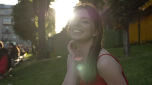 Joven mujer bonita con el pelo de jengibre tocando la cabeza y mirando a la cámara, sentado en la hierba en la calle — Vídeo de stock