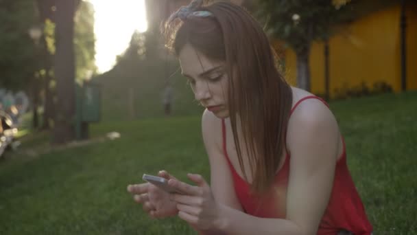 Молодая красивая женщина с рыжими волосами болтает по телефону и сидит на траве на улице — стоковое видео