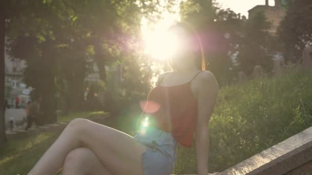 Young charming woman with ginger hair sitting on grass on street and waving hair, sun shining brightly — Stock Video