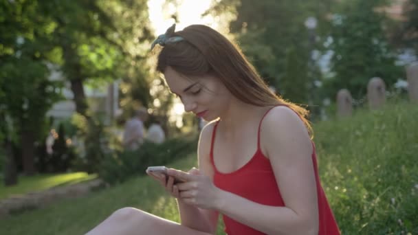 Giovane bella donna con i capelli rossicci chatta al telefono e seduta sull'erba nel parco — Video Stock