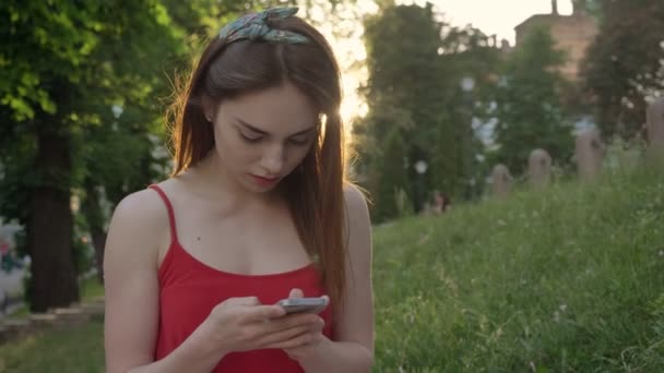 Charmante jongedame met gember haar texting op telefoon en zittend op het gras in park, glimlachend — Stockvideo