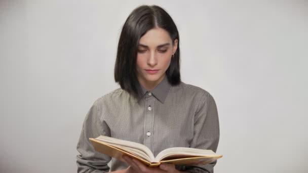 Jeune jolie femme aux cheveux bruns courts tenant livre et lecture, puis regardant à la caméra et souriant, fond blanc — Video