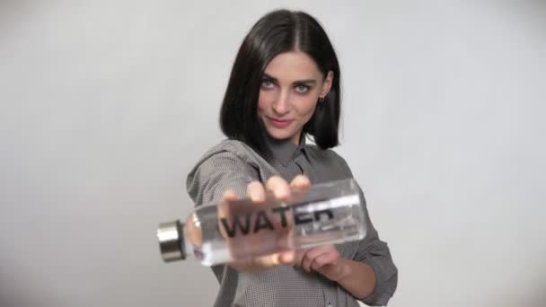 Jonge mooie vrouw met kort bruin haar houden en wijzen fles met water in de camera, tonen van grote duim, gelukkig, witte achtergrond — Stockvideo