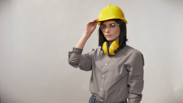 Mladý tvůrce ženy stojící v sluchátka, brýle a helmu dotýká helma, vážný výraz, bílé pozadí — Stock video