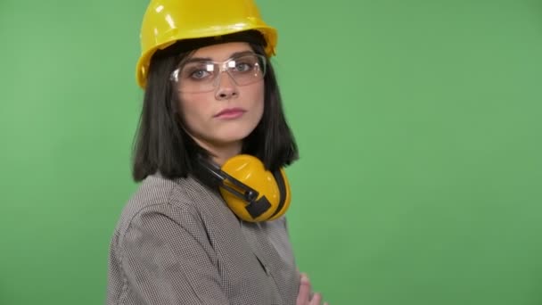 Mujer joven constructora de pie en auriculares, gafas y casco con las manos cruzadas mirando a la cámara, parpadeando, croma clave de fondo — Vídeo de stock