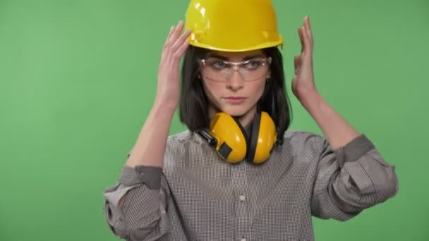 Jovem mulher bonita construtor de pé em fones de ouvido, óculos e capacete fixando seu capacete, sério, croma fundo chave — Vídeo de Stock