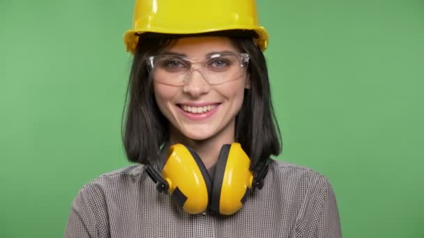 Joven hermosa constructora mujer de pie en auriculares, gafas y casco mirando en la cámara y posando, sonriente, croma clave de fondo — Vídeos de Stock