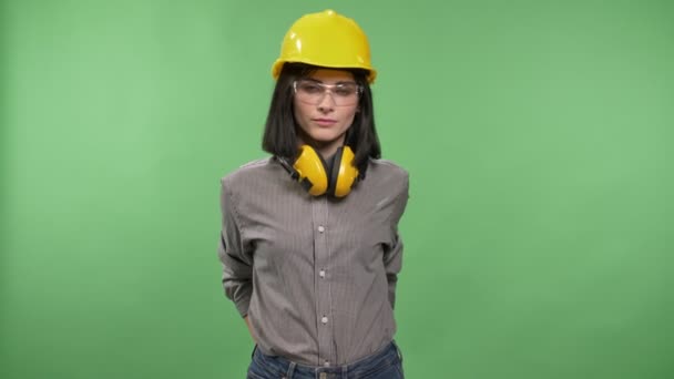 Молодая красивая женщина-строитель, стоящая в наушниках, очках и шлеме с руками в карманах и глядя в камеру, хрома ключ фон — стоковое видео