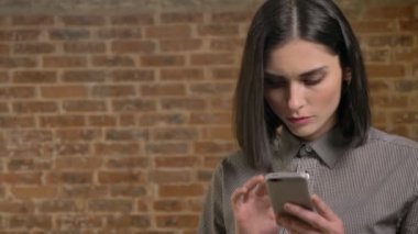 Telefona yazma ve konsantre, onun dudak ısırma genç güzel kadın tuğla duvar arka plan portresi